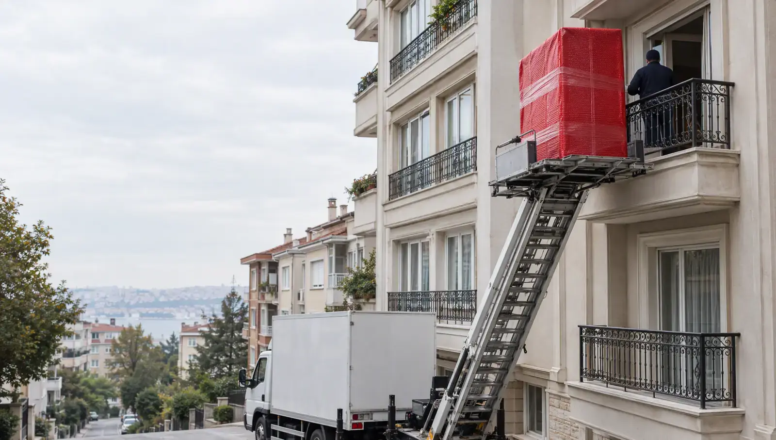 Dar bir İstanbul sokağında dördüncü kat balkonuna uzanan dış cephe asansörü; platformda kırmızı ambalajlı gardırop